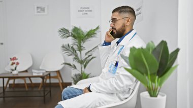 Beyaz laboratuvar önlüklü sakallı genç bir adam lobide oturup telefonla konuşuyor..