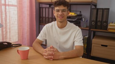 Genç, yakışıklı, İspanyol bir adam, düzenli dosyalar ve kitaplarla çevrili bir fincan kahveyle birlikte, bir ofisin içinde güven içinde oturuyor..