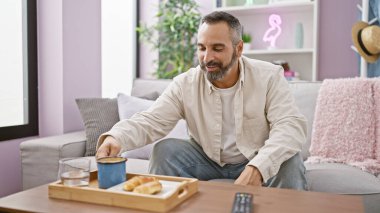 Olgun sakallı bir adam, rahat bir oturma odasındaki kanepede kahvaltıdan zevk alır.