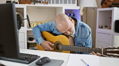 Kel bir adam, müzik aletleri ve kayıt ekipmanlarıyla çevrili bir ofiste gitar çalıyor..