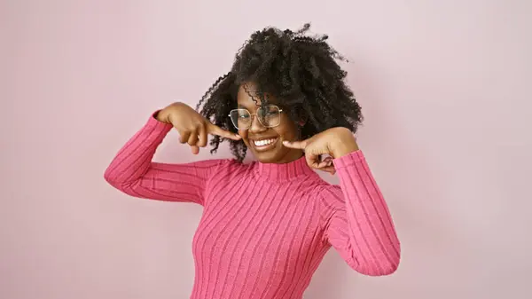 stock image A happy black woman with glasses pointing to her smile indoors against a pink background