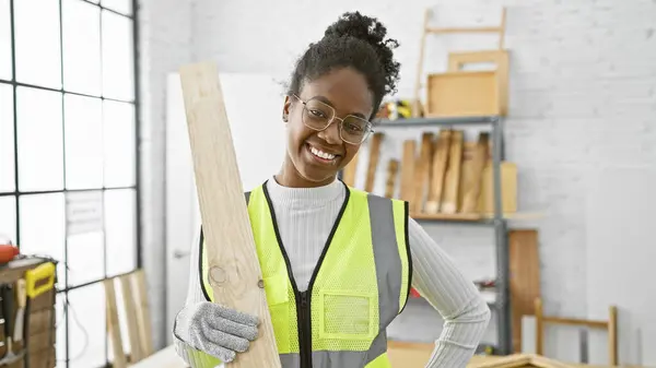 stock image A smiling black woman wearing safety goggles and gloves holds lumber in a bright carpentry workshop.