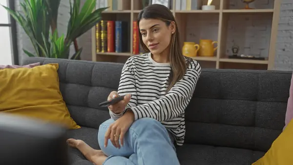 Stock image A young hispanic woman in casual clothing relaxes on a sofa at home, holding a remote control