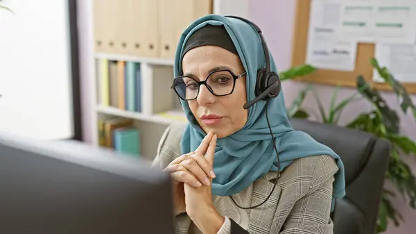 stock image A thoughtful middle-aged woman wearing a headset and glasses in an office, symbolizing professional customer service.