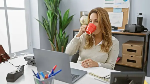 Stock image Hispanic woman drinking coffee at her modern office desk indoors while working on laptop.