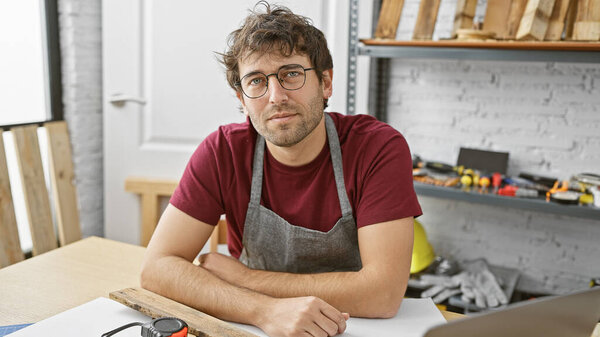 A young hispanic man with a beard poses in a carpentry workshop wearing an apron.
