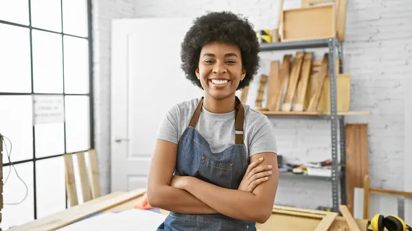 stock image Smiling black woman in apron standing with arms crossed in a bright carpentry workshop.