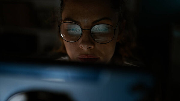 Сосредоточенная молодая женщина в очках, освещенная светом экрана ноутбука, работая допоздна в темной офисной комнате.