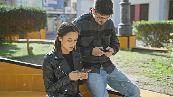 stock image A man and a woman focused on smartphones while sitting together on a bench in a sunny urban setting
