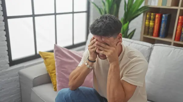 stock image A distressed hispanic man covering his face with hands while sitting on a sofa indoors.