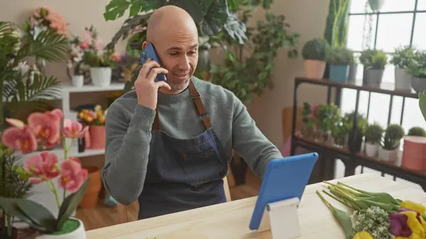 stock image A bald hispanic man with a beard talks on a phone while browsing a tablet in an indoor flower shop.