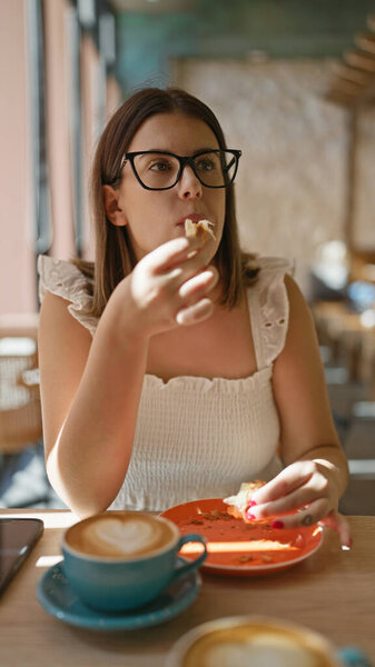 Portrait of a beautiful hispanic woman enjoying a delicious croissant. see this brunette in glasses savouring this sweet meal at a sunny cafeteria table, making breakfast an indoor delight.