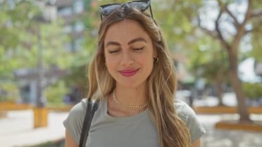 Güneşli bir şehir parkında kameraya gülümseyen güneş gözlüklü genç bir kadın.