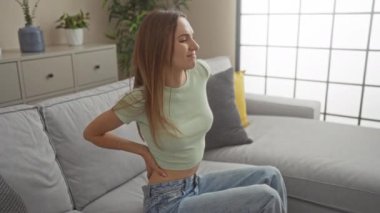 Genç, çekici, sarışın bir kadın modern bir oturma odasında modern bir dekorasyonla oturuyor. Gri bir kanepede sırt ağrısı ve rahatsızlık yaşıyor. Etrafı ev bitkileri ve yumuşak yastıklarla çevrili..