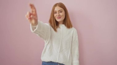 Neşeli, genç, kızıl saçlı, kendine güvenen, pozitif zafer işaretinin üzerinde duran bir kadın. İki parmağında V işareti var, pembe arka planda kameraya gülümsüyor..