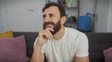Evde orta yaşlı bir adam, eli çenesinde, düşünceli bir bakışla sorguluyor. Beyaz tişört giyiyor, düşünceli ifadesi içsel şüpheleri dile getiriyor..