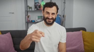 Neşeli orta yaşlı bir adam, dost canlısı bir gülümseme takınıyor, telefonla jest yapıyor, parmaklarını kaldırmış, rahat evinde telefon taklidi yapıyor.