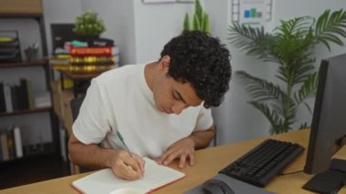 Ofiste çalışan genç bir adam bilgisayarlı bir masada oturup kahve içerken not defterine not alıyor. Etrafı kitaplarla ve bitkilerle çevrili profesyonel bir çevreyle çevrili.