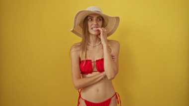 Bikinili ve yaz şapkalı kadın endişeli görünüyor ve stres ve stres yüzünden tırnaklarını yiyor sarı arka planda izole edilmiş ve elleri sorun çıkarıyor.