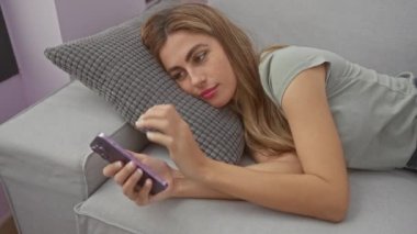 Genç sarışın bir kadın akıllı telefonuyla evinde kanepede uzanıyor, rahatlamış, sıkılmış ve sıradan bir hayat tarzında esniyor..