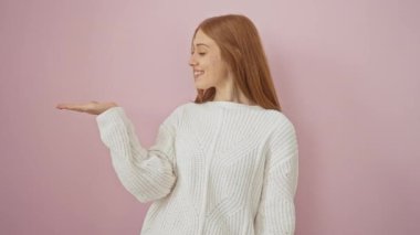 Süveteri içinde ışıl ışıl parlayan neşeli kızıl saçlı bir kadın, hoş geldin jesti yapıyor, ürününüzü pembe, şık bir arka plana karşı sıcak bir gülümsemeyle sunuyor..