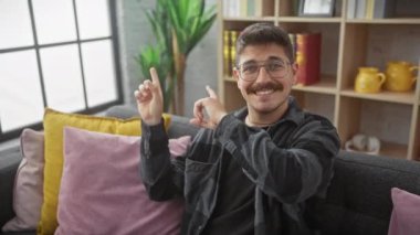 Evdeki kanepede otururken mutlu bir şekilde yan tarafı gösteren, gözlüklü, neşeli, genç İspanyol bir adam. Kendine güveni tam bir gülümsemeyle kameraya bakıyor..