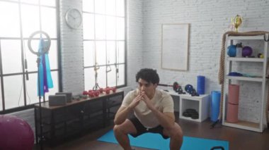 İspanyol adam, çeşitli spor aletleri ve aksesuarlarla çevrili, iyi donanımlı bir spor salonunda mavi bir paspasa çömeliyor..