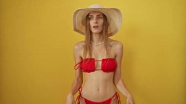 Yaz bikinili bir kadın şapkasını kaldırmış, sarı arka planda şaşkın şaşkın bakıyor mutlu, şaşırmış, beyaz bir kadın ayakta ve gülümsüyor.
