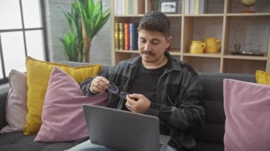 İspanyol adam evde çalışırken gözlük temizliyor, renkli yastıklı rahat ve şık bir oturma odası gösteriyor..