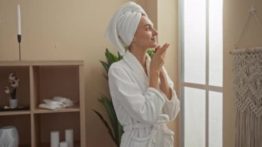 Kadın, sağlık merkezindeki spa deneyimini beyaz bornoz ve kafasında havluyla pencereden dışarı bakarak rahatlatıyor.