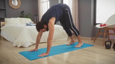 Sakallı bir adam yatak odasındaki mavi bir paspasın üzerinde yoga yapıyor. Bu da içeride sağlıklı bir yaşam tarzını gösteriyor..