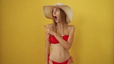 Bikinili ve yaz şapkalı kadın şaşırmış bir ifadeyle işaret ediyor, ağzı açık şaşırmış, izole edilmiş sarı arka planda, beyaz kadın neşeli bir şekilde parmaklarıyla kenarı işaret ediyor.