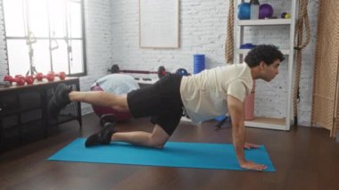 İyi donanımlı bir spor salonunda mavi bir paspasın üzerinde egzersiz yapan genç bir İspanyol adam çeşitli spor aletleriyle kendini adamış ve gücünü açık, düzenli bir iç mekan ortamında sergiliyor..