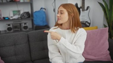Kendine güvenle dolup taşan genç, neşeli kızıl saçlı kadın sıcak evinin kenarını işaret ederek mutlu bir şekilde yüzünü kaplayan samimi bir gülümseme.