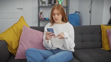 Neşeli, kızıl saçlı bir kadın, oturma odasındaki gri koltukta akıllı telefon kullanıyor..