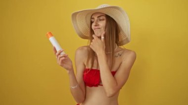 Bikinili bir kadın elinde güneş kremi tutarken ciddi bir yüz, çenesinde el, sarı arka planda izole edilmiş düşünceli bir fikir olduğunu düşünüyor.