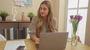 Sarışın genç bir kadın, iyi ışıklandırılmış bir ofis ortamında akıllı telefon ve laptop kullanırken gülümsüyor..