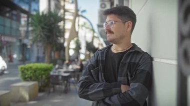 Güneşli bir şehir sokağında kolları çapraz duran bıyıklı neşeli, genç bir İspanyol..