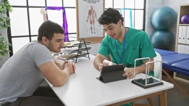 Fizyoterapi kliniğinde erkek hastayla birlikte tablet kullanan önlüklü bir terapist.