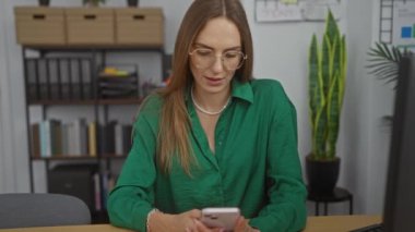 Ofis ortamında genç sarışın bir kadın gözlüklü ve yeşil gömlekli, kitaplık ve ev bitkileriyle çevrili akıllı telefonuna sevinçle bakıyor..