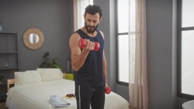 Olgun sakallı bir adam modern bir yatak odasında dambıllarıyla egzersiz yapıyor, evde spor yapmaya odaklanıyor..
