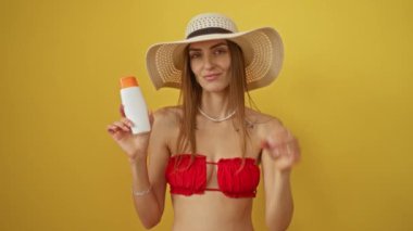Bikinili kadın elinde güneş kremi tutarak işaret veriyor, mutlu ve gülümsüyor, izole edilmiş sarı arka planda parmaklarının arasından bakıyor.