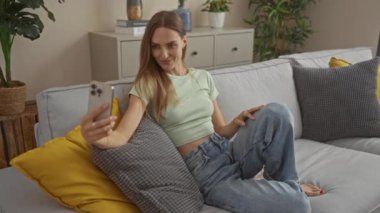 Genç, çekici, sarışın bir kadın, modern mobilya ve bitkilerle süslenmiş rahat bir oturma odasında uzanırken selfie çekiyor..