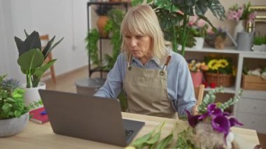 Bej önlüklü sarışın bir kadın canlı bir çiçekçide, ahşap bir masada dizüstü bilgisayar üzerinde çalışırken çiçekleri düzenliyor..
