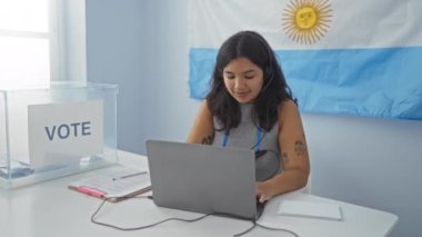 Çekici, genç bir İspanyol kadın kapalı bir üniversite odasında dizüstü bilgisayar üzerinde çalışıyor. Arjantin bayrağı ve arka planda şeffaf bir oy sandığı var..