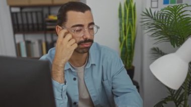 Çiçekler ve kitaplıklarla çevrili bir ofiste çalışan bıyıklı İspanyol bir adam.