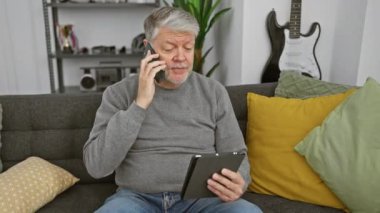 Son sınıf öğrencisi elinde tablet, modern bir oturma odasındaki kanepede otururken akıllı telefonuyla konuşur..