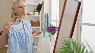 Düşünceli sarışın bir kadın, parlak bir odada çiçekli giysileri ve kıyafet seçimlerini yansıtan bir aynayı inceliyor..