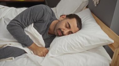 Genç adam yatakta uzanıyor, rahat bir yatak odasının içindeki beyaz yastıkların üzerinde huzur içinde yatıyor, gri bir uyku giysisi giyiyor, evinde huzurlu ve huzurlu bir atmosferi çağrıştırıyor.