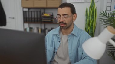 Bıyıklı bir adam ofiste çalışıyor, gözlük takıyor, stresli hissediyor, etrafı kapalı bitkiler ve kitaplıklarla çevrili.